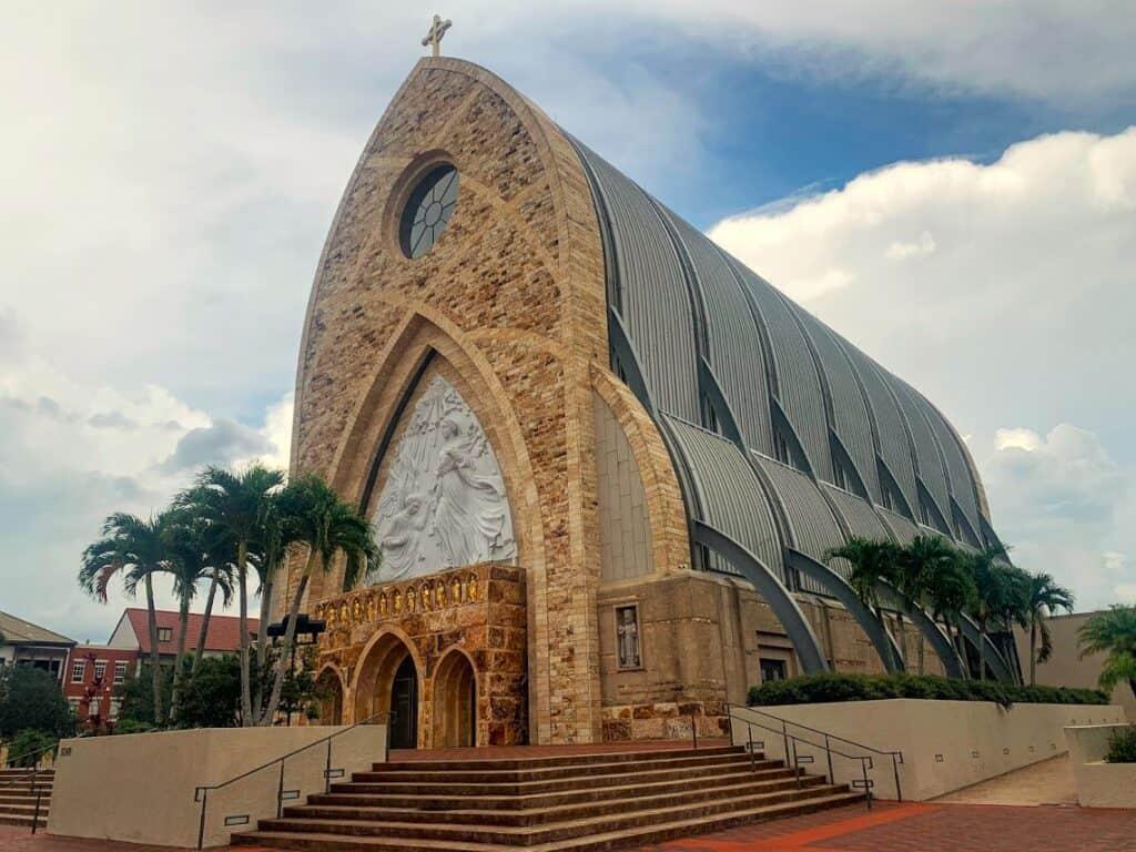 The Ave Maria Oratory, a stone church with a soaring arched facade, framed by palm trees in Ave Maria, Florida.